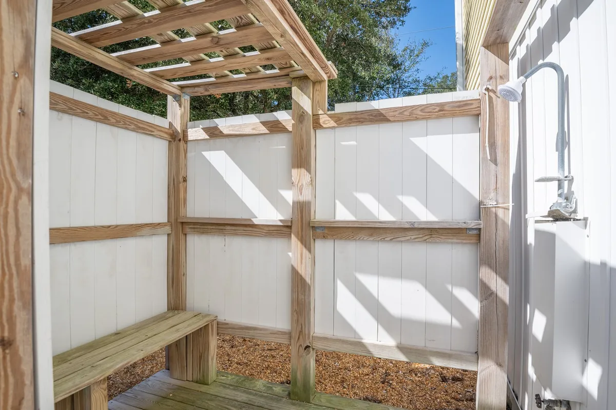 A refreshing outdoor shower area with a wooden bench, sturdy shower fixture, and a pergola roof allowing dappled sunlight. The setup provides a tranquil spot to rinse off after a beach day, surrounded by natural elements for a serene, quintessential coastal experience.