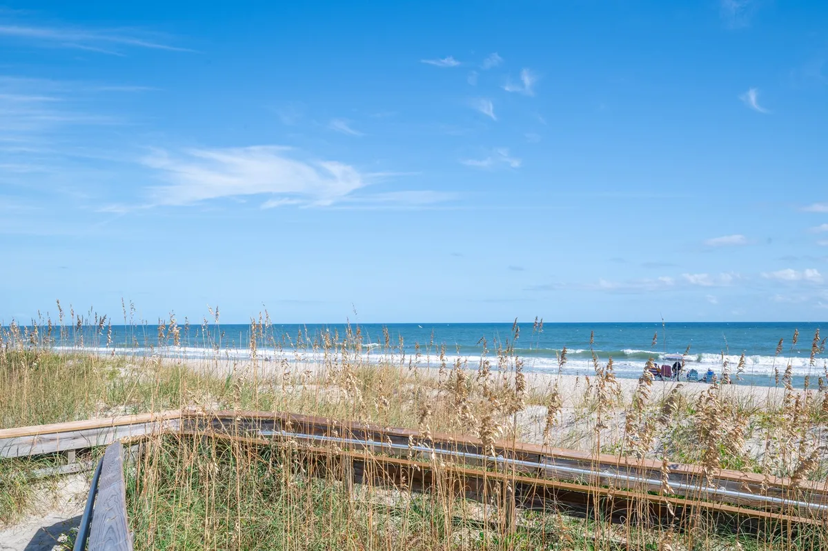 The image showcases a tranquil stretch of Kure Beach with soft dunes, the expansive ocean, and a blue sky overhead. People can be seen sunbathing, walking, and enjoying the peaceful shoreline, evoking a sense of relaxation and simple coastal pleasures.