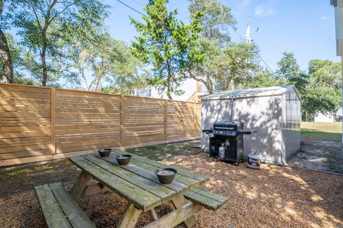This image features a wooden picnic table and propane grill set on a grassy area, bordered by a wooden fence and with a small shed nearby. The scene exudes a welcoming and relaxed vibe, perfect for outdoor dining or gathering with others. The setting shows ample space for both people and pets to enjoy the backyard.