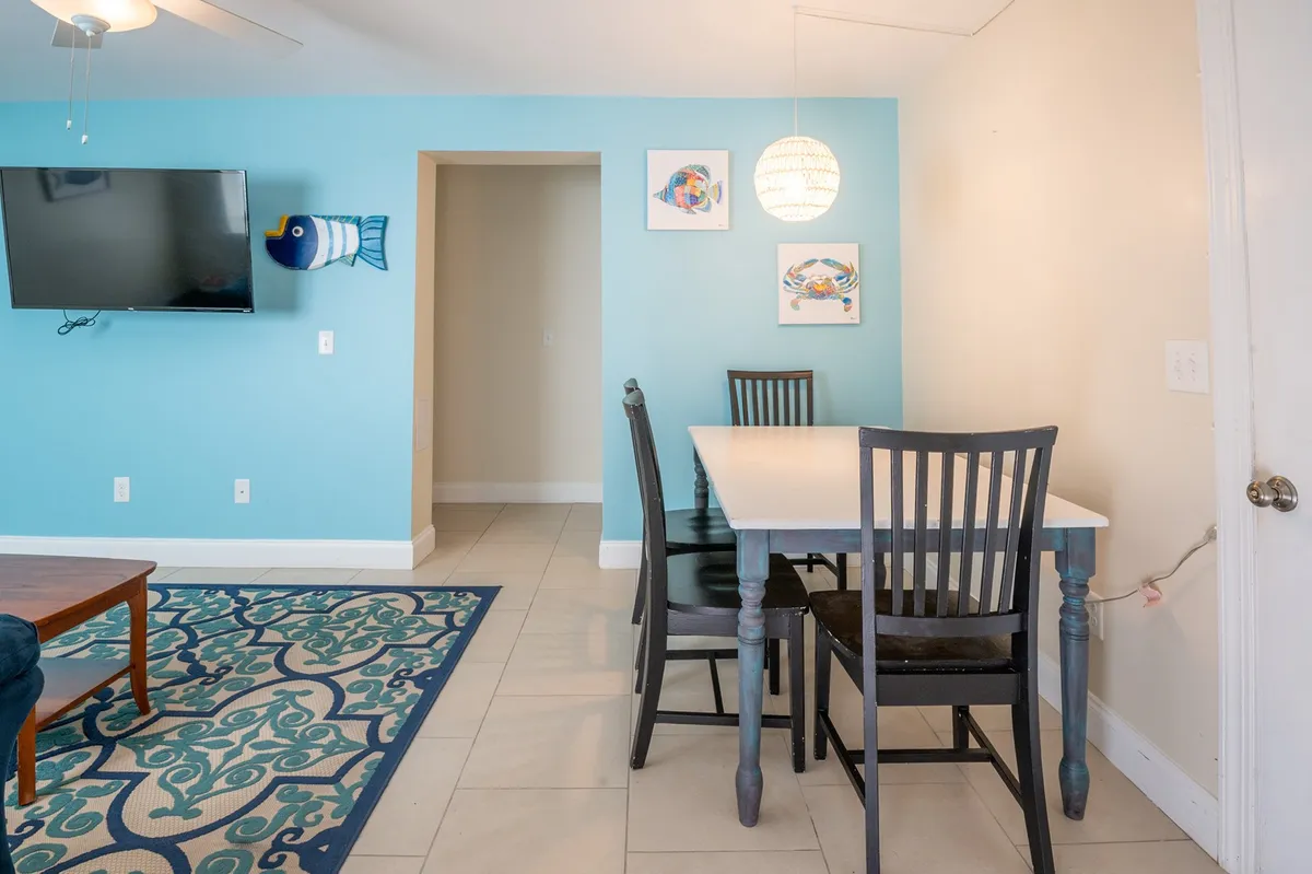 A casual, inviting dining area featuring a modest table set up for shared meals, a television for entertainment, tasteful wall art, and a cozy blue accent wall with a colorful rug underfoot. The space is designed for relaxed family gatherings, casual takeout dinners, or a quick breakfast from the coffee bar.