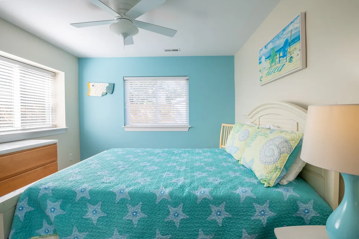 A bright queen bedroom with a bed dressed in a star-patterned quilt, colorful wall art, a window with blinds, and a nightstand with a lamp. The space feels peaceful and inviting, bathed in natural light—ideal for relaxation.
