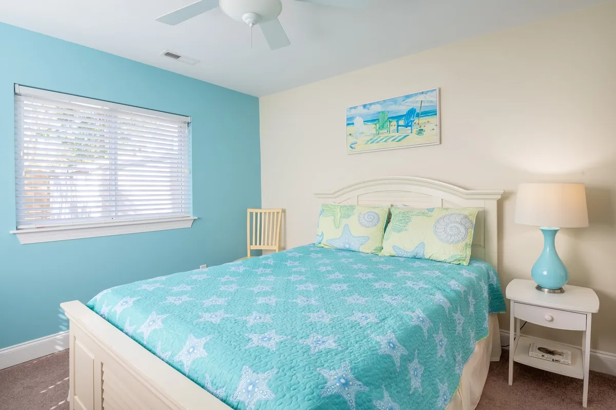 A tranquil bedroom with a bed made up in a starfish-patterned quilt, decorative pillows, a turquoise-base lamp, and tasteful wall art. The soft sunlight and cozy setup evoke a sense of calm and relaxation.