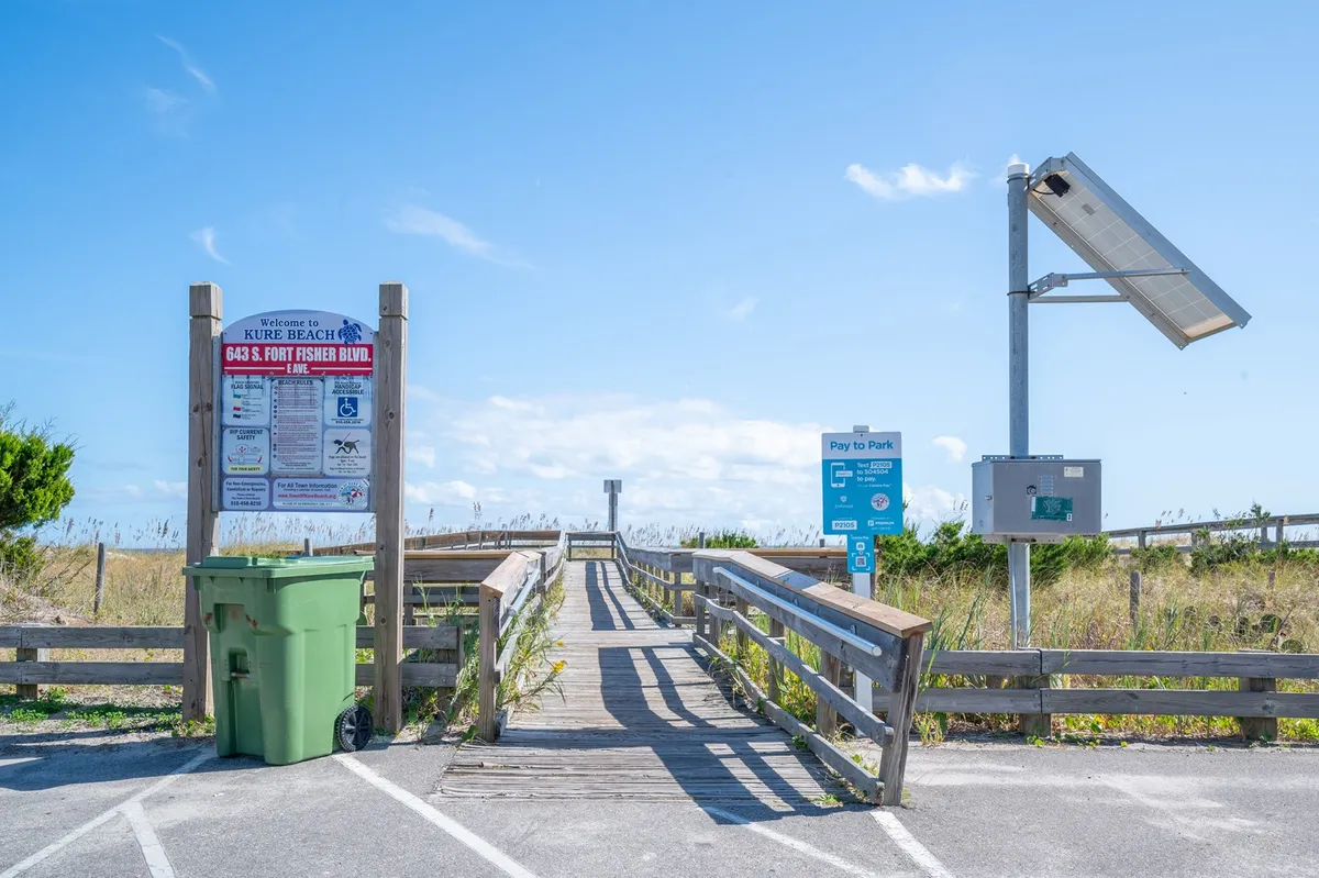 A sandy wooden walkway leads directly to the beach, marked by a welcoming beach access sign. The surrounding area features dunes and a few coastal touches such as a green trash can and a solar panel, capturing the relaxed vibe of an easy stroll to the shoreline.