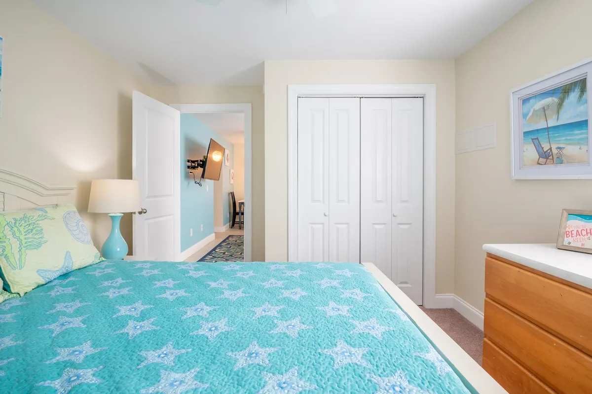 A cozy queen bedroom featuring a bed with a star-patterned quilt, a nightstand with a lamp, a chest of drawers, and framed beach-themed art. Soft natural light fills the room, creating an inviting and tranquil atmosphere ideal for rest.